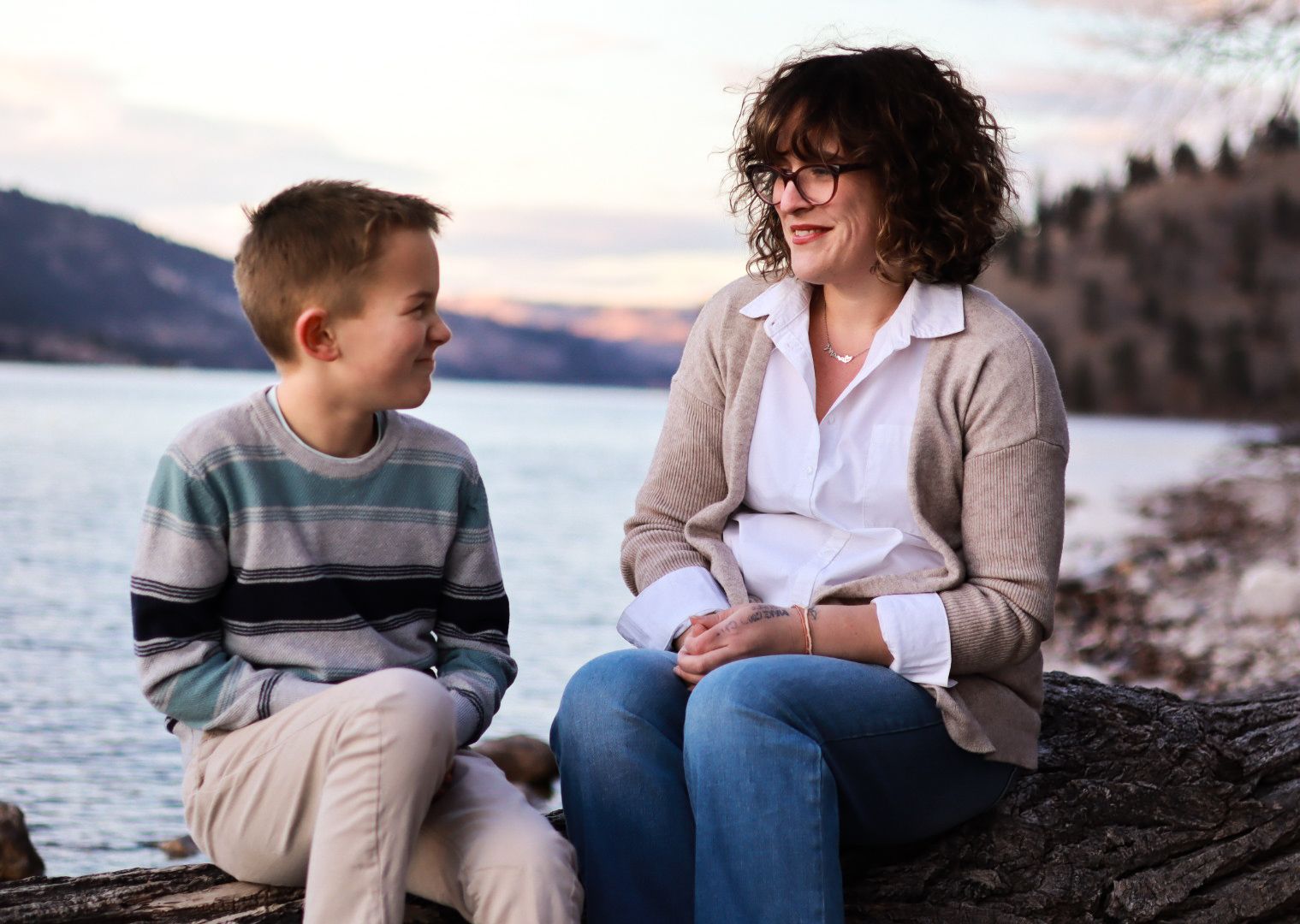 Parent and child share a tender moment while sitting by a lake at sunset against a mountain backdrop.