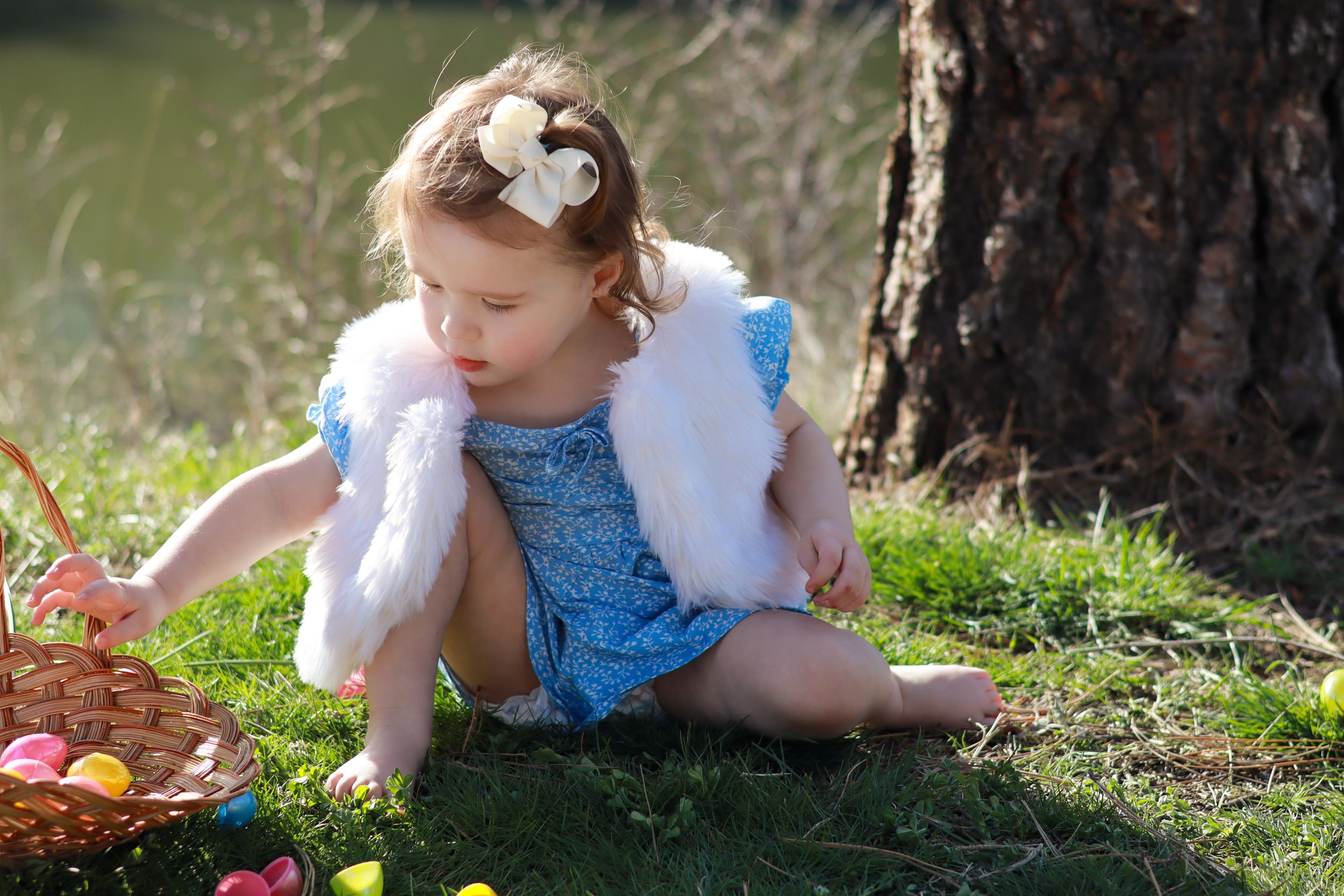 A small figure in a blue dress and white shawl sits on mossy ground near autumn leaves and a tree trunk.