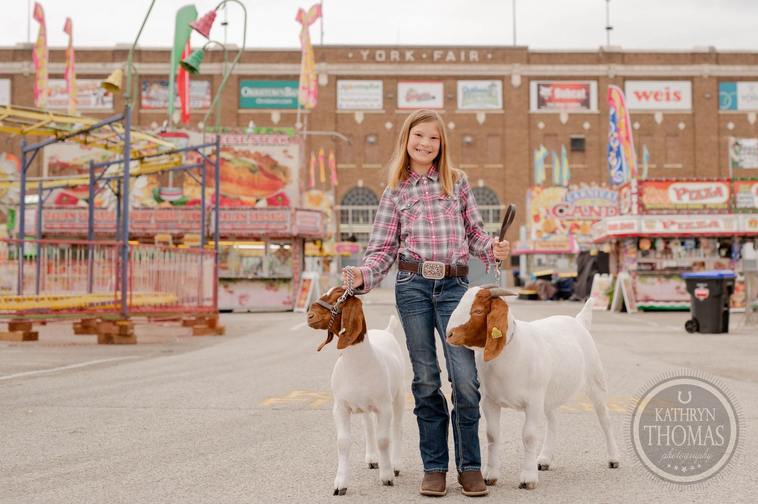 Two goats stand on leashes in front of York Fair building with carnival rides visible.