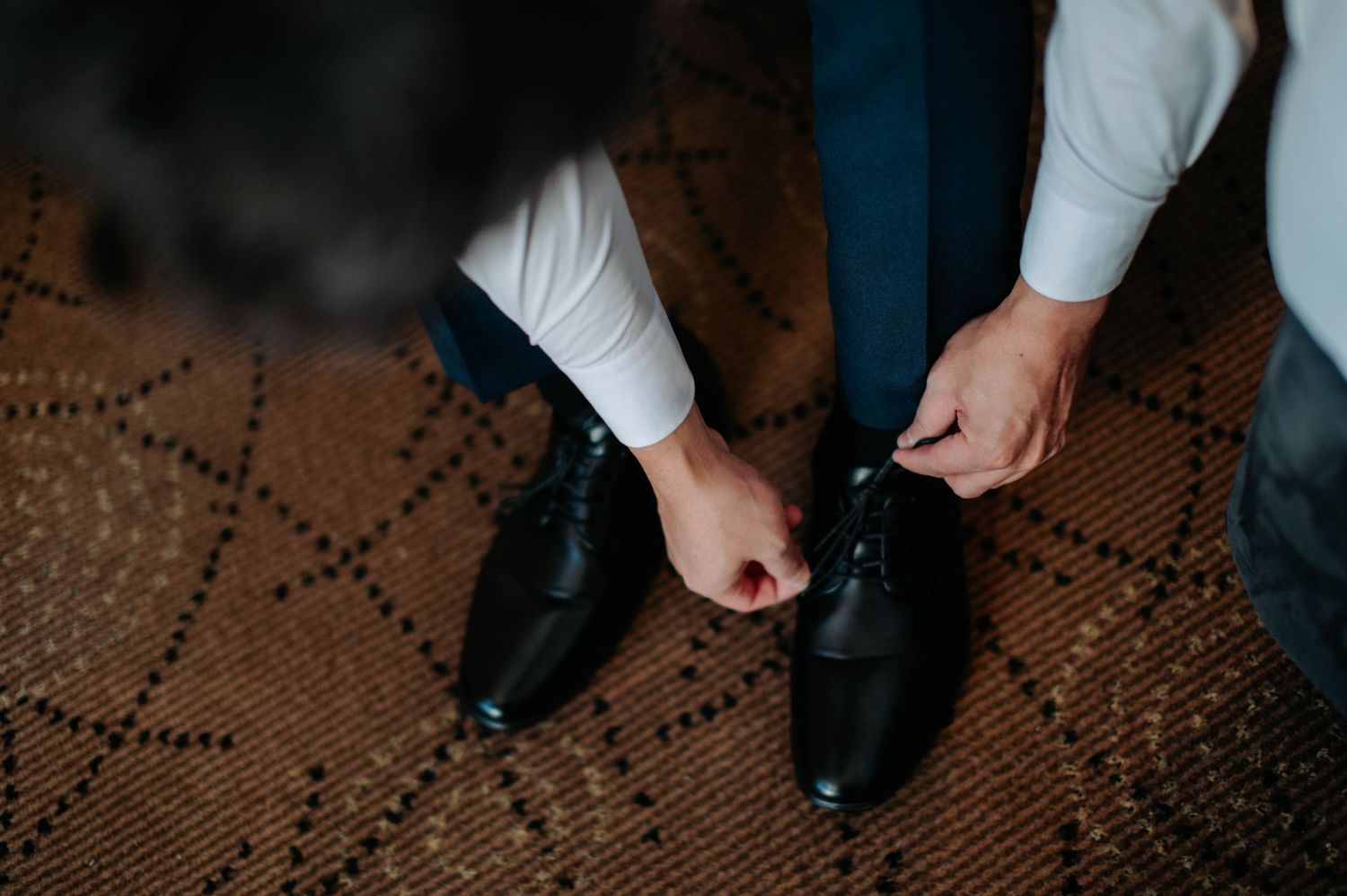 Close-up shot of hands tying formal black leather dress shoes on patterned carpet.