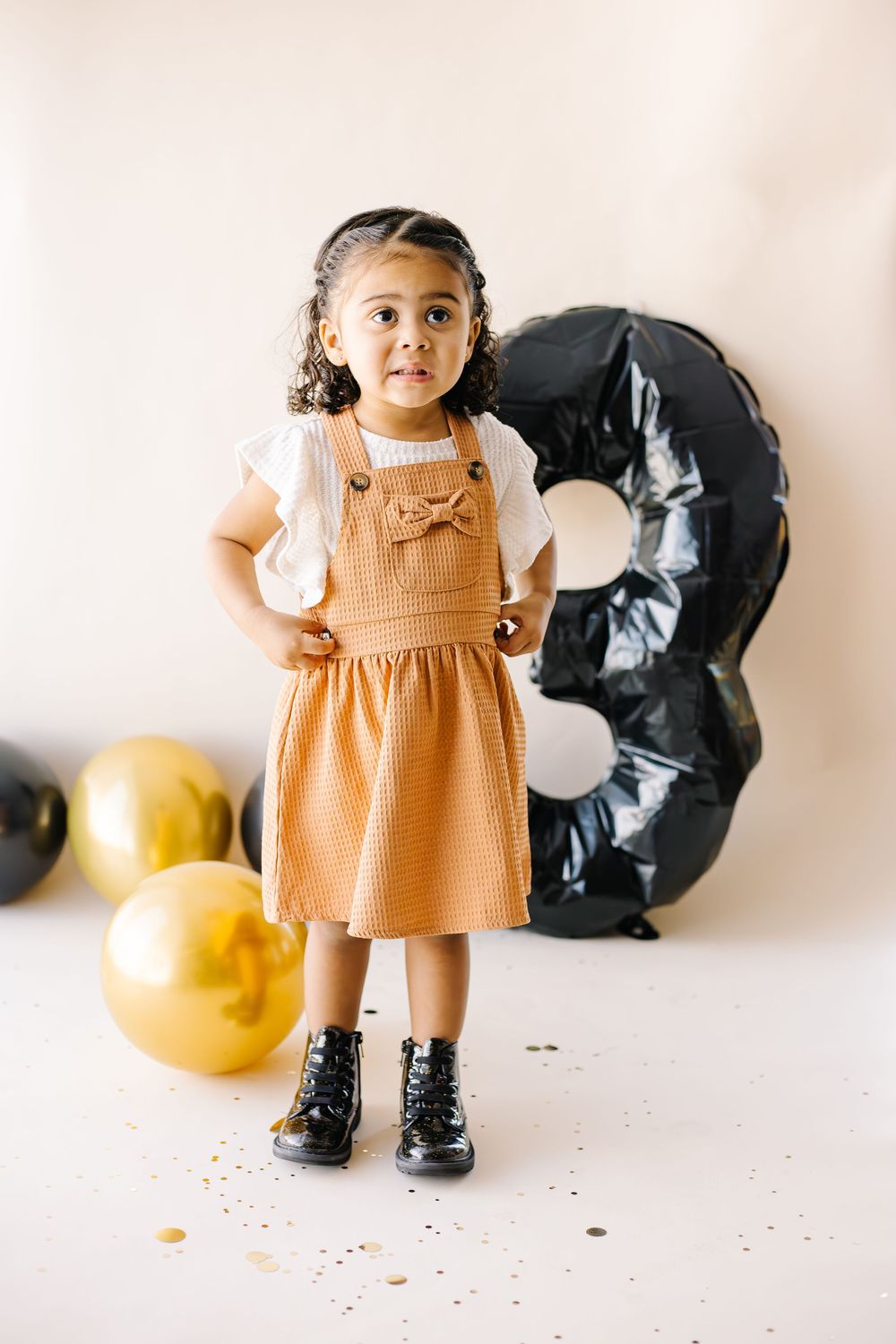 A toddler in a tan pinafore dress and black boots stands near black and gold balloon decorations.