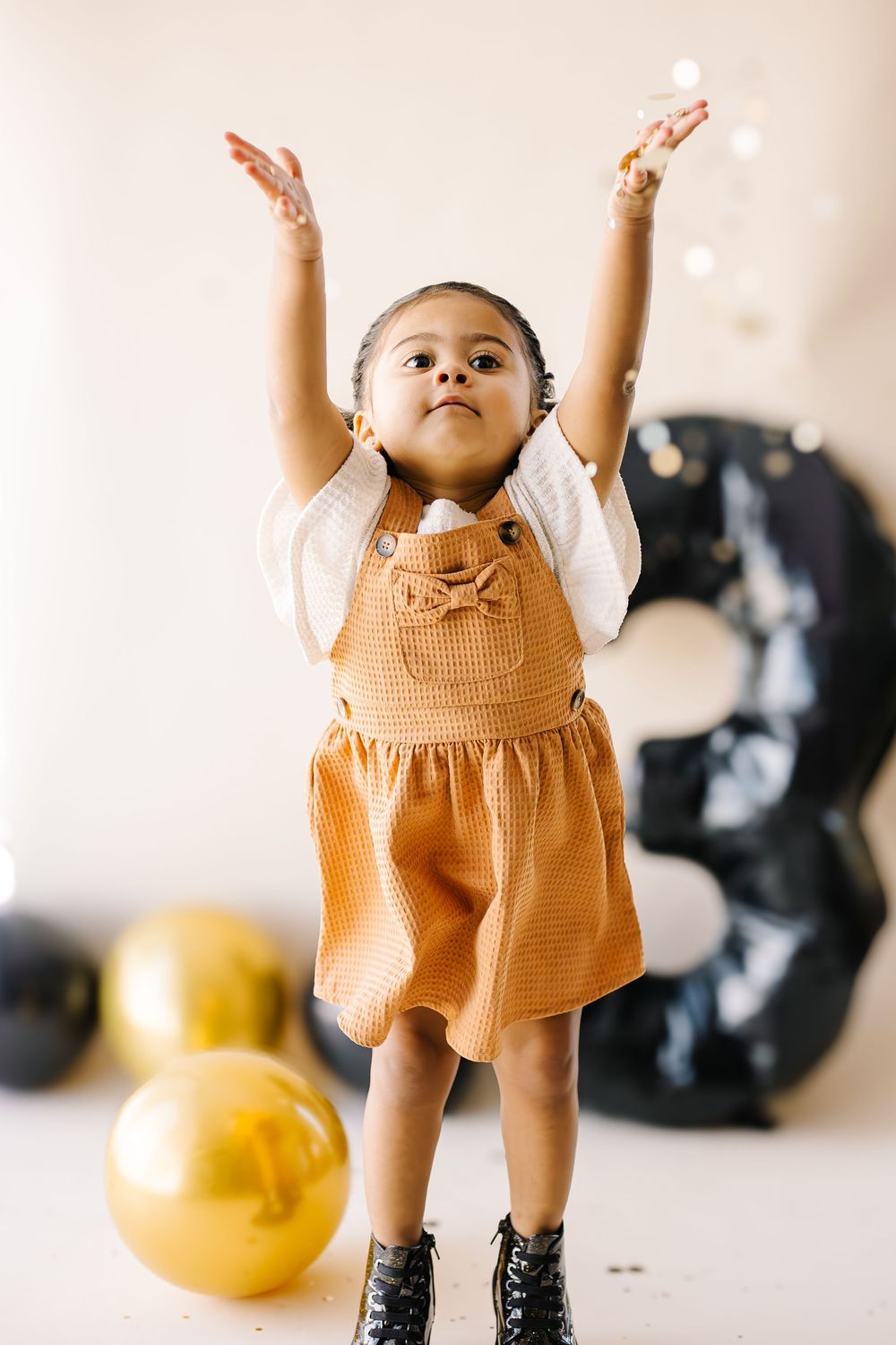 Young child wearing orange dress reaching upward with both arms near yellow balloons and black decor.