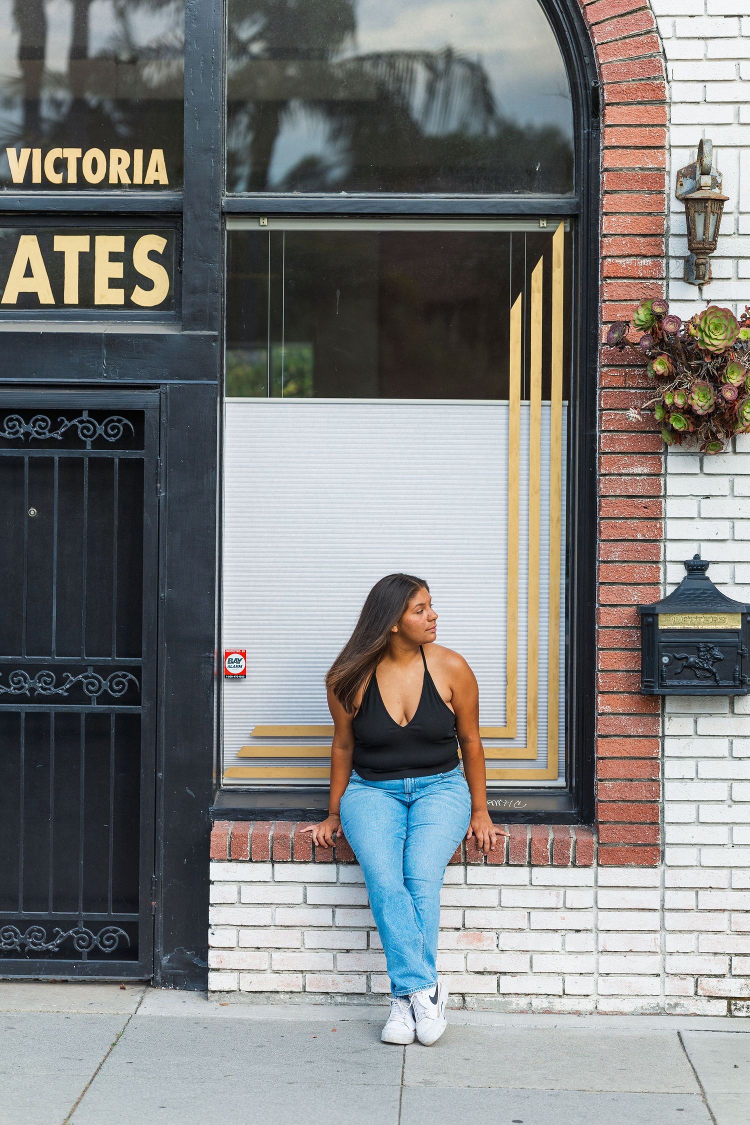 A person in black tank top and blue jeans leans against a white brick wall near a storefront with Victoria signage.
