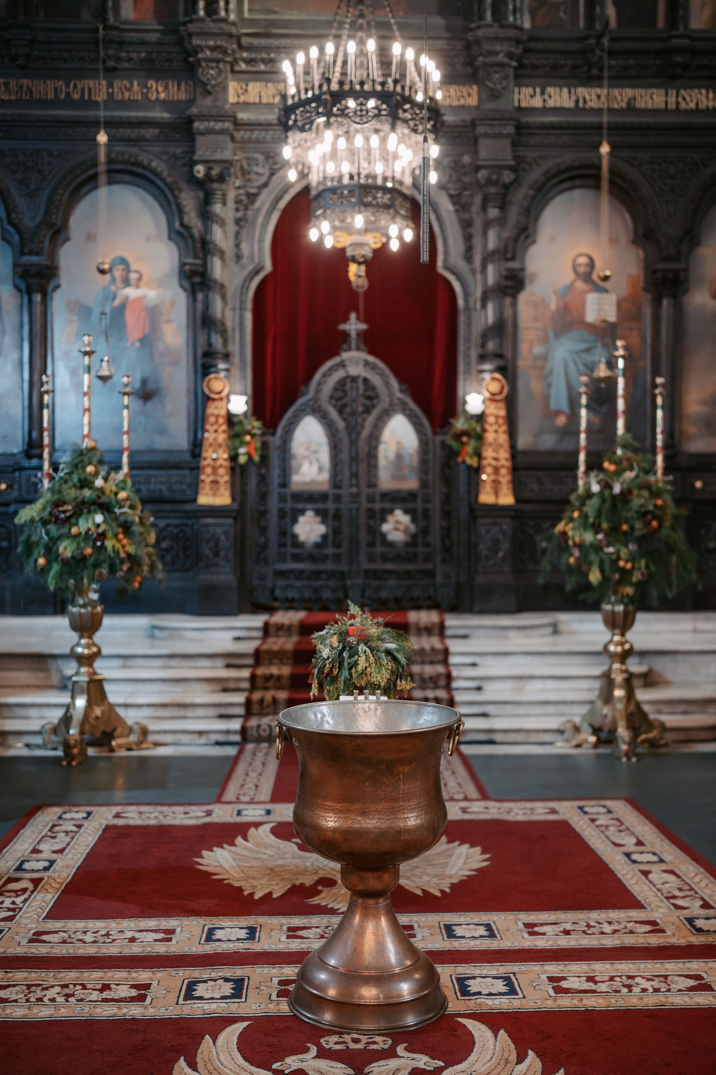 Interior of an ornate Orthodox church with chandelier, red carpet, and decorative holy water font in the foreground.