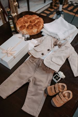 White baby clothes outfit with brown shoes and wrapped gift box displayed on dark wooden surface.