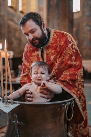 Orthodox baptism ceremony showing a priest in red vestments performing the sacrament over a baptismal font.
