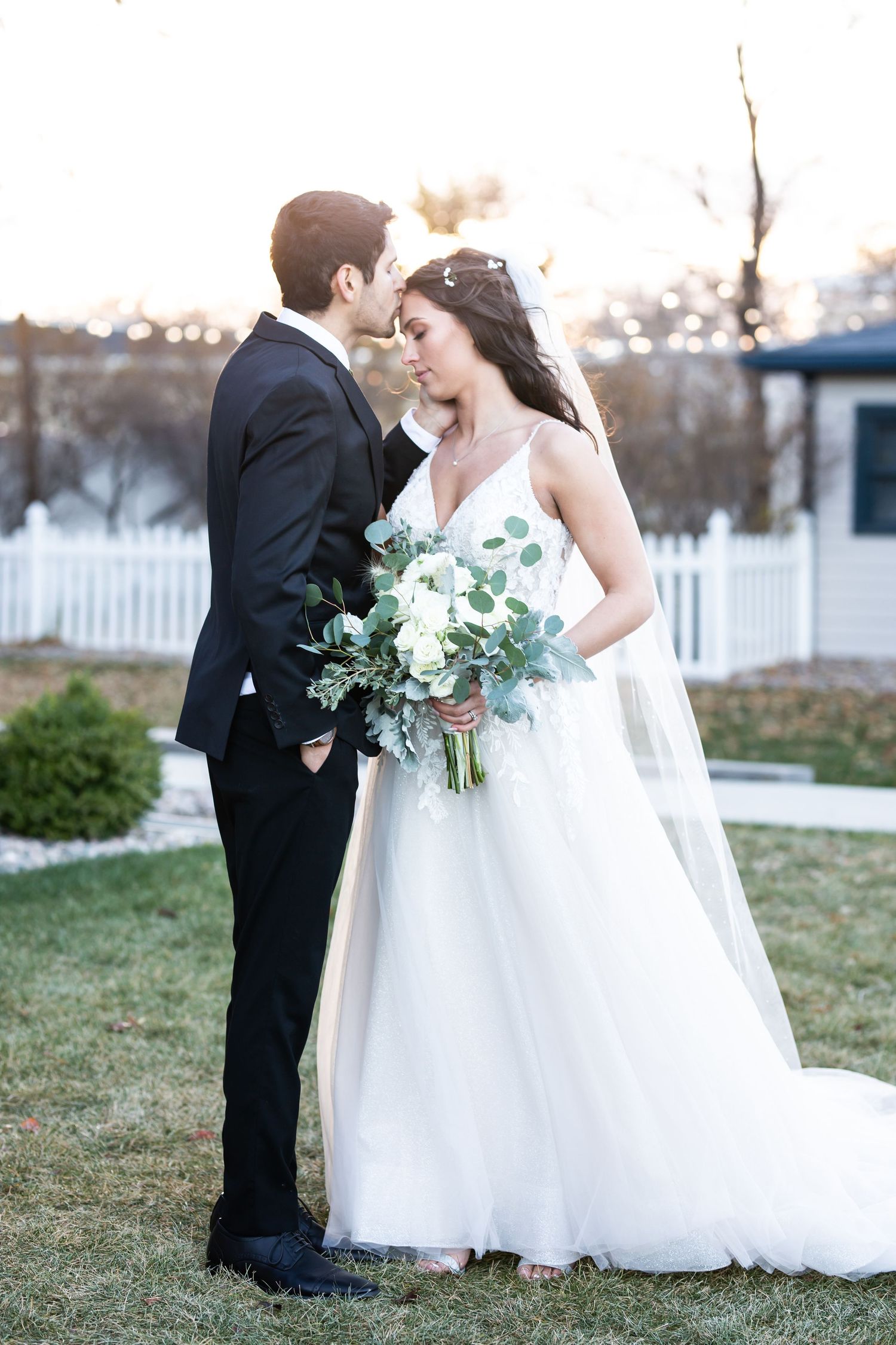 A bride in a flowing white gown holds a eucalyptus bouquet while sharing an intimate moment with her groom outdoors.