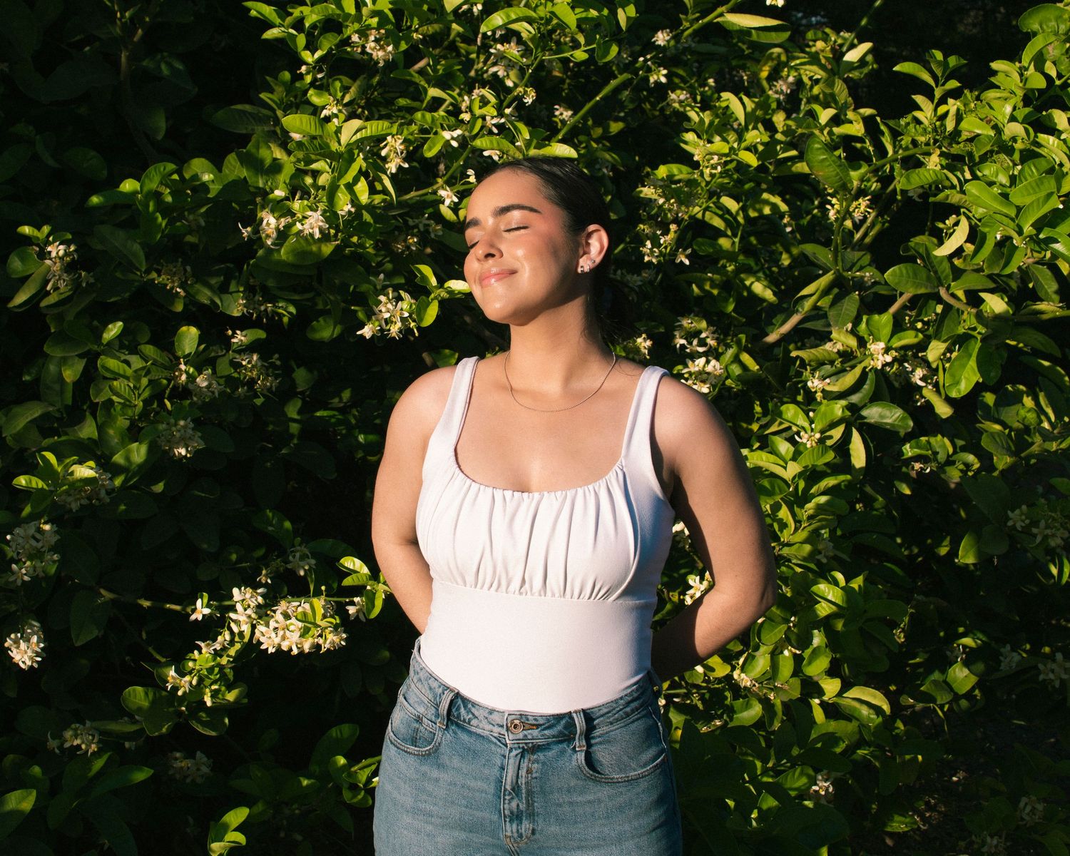 Young woman in a white tank top and jeans stands in front of lush green foliage on a sunny afternoon, basking in the sunlight.