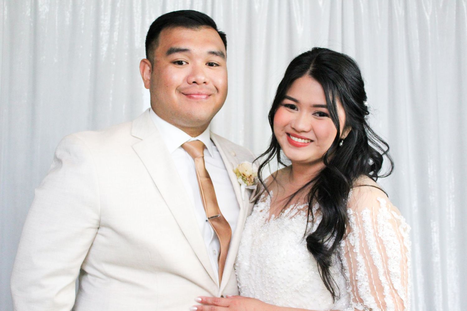 A smiling couple poses together in formal white attire during what appears to be a wedding portrait session.