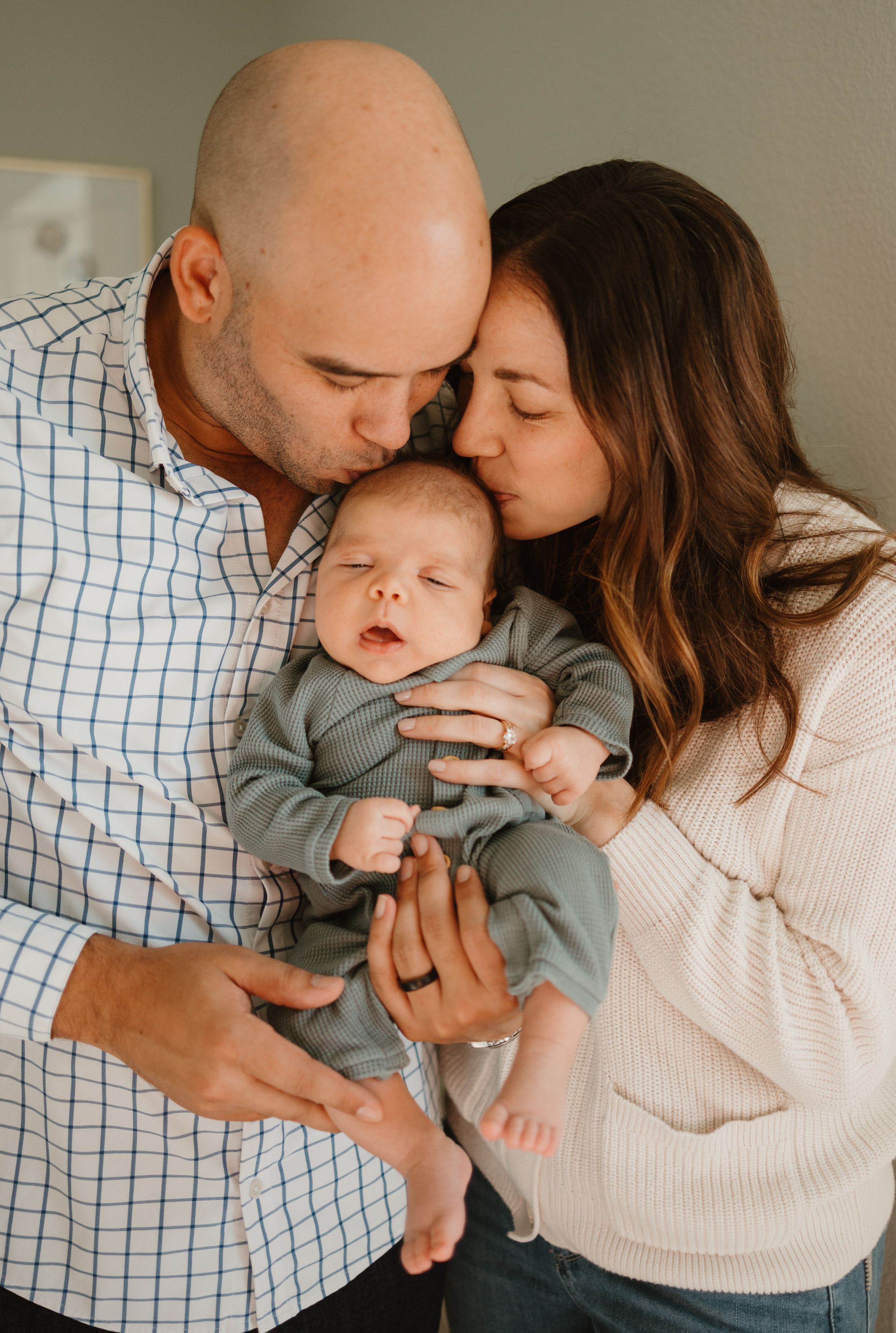 An intimate family portrait shows parents sharing a tender moment with their sleeping newborn baby in gray clothing.