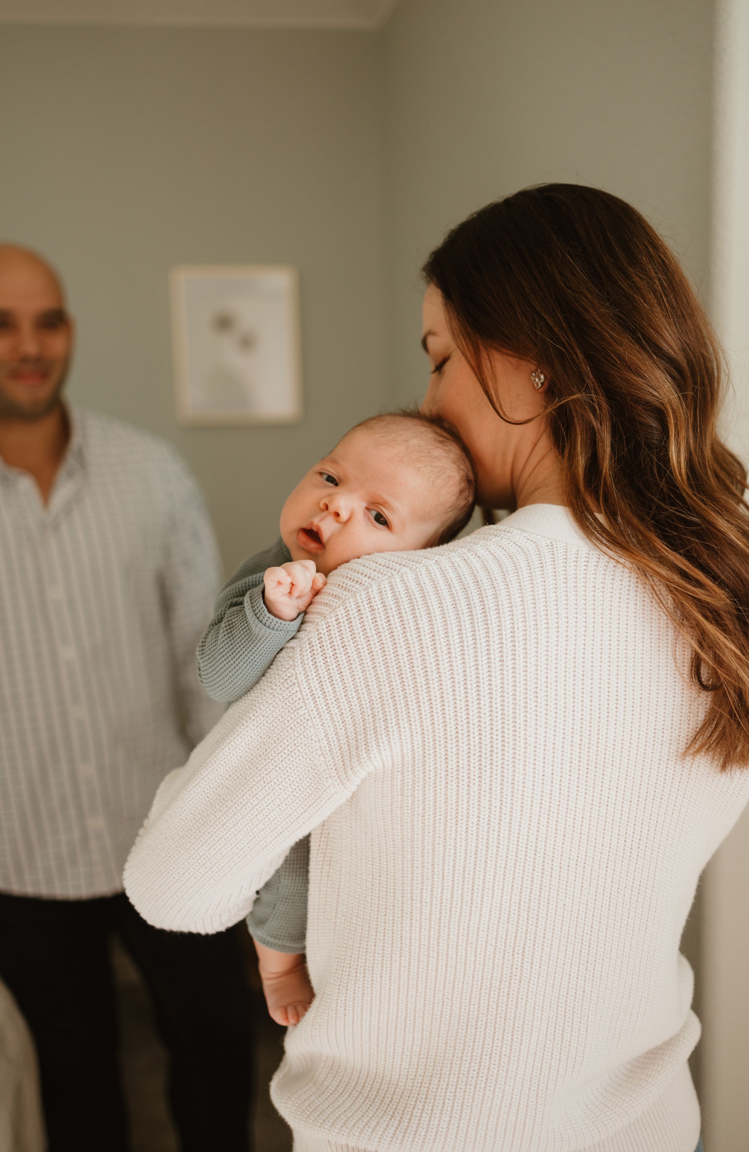 A parent in a white sweater holds a sleeping baby while another adult stands in the background of a peaceful nursery scene.