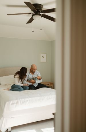 A couple sits together on a white bed under a ceiling fan in a bright minimalist bedroom.