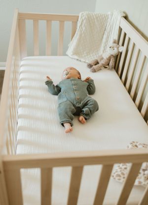 A baby wearing a teal onesie sleeps peacefully in a white wooden crib with a plush toy.