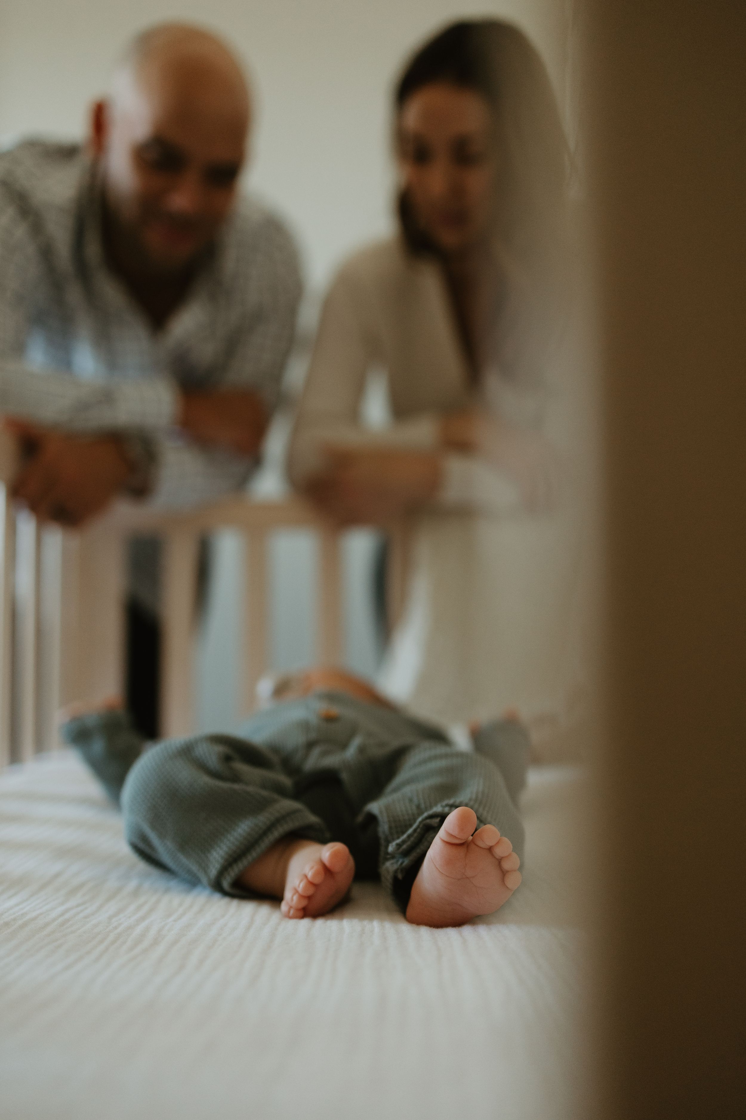 Small baby feet in gray clothes lying on white bedding while parents lovingly watch from behind.