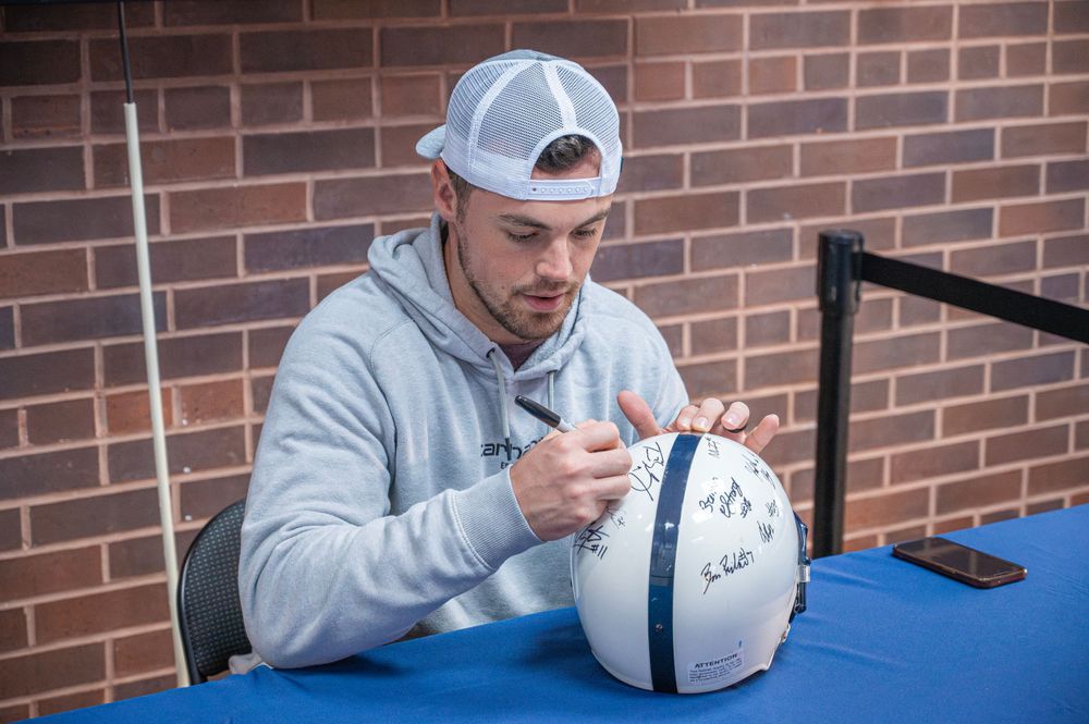 Someone in a gray hoodie and baseball cap signs a football while seated at a table against a brick wall backdrop.