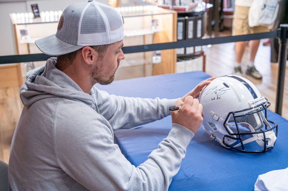 A person in a grey hoodie and baseball cap signs a white football helmet on a blue surface.