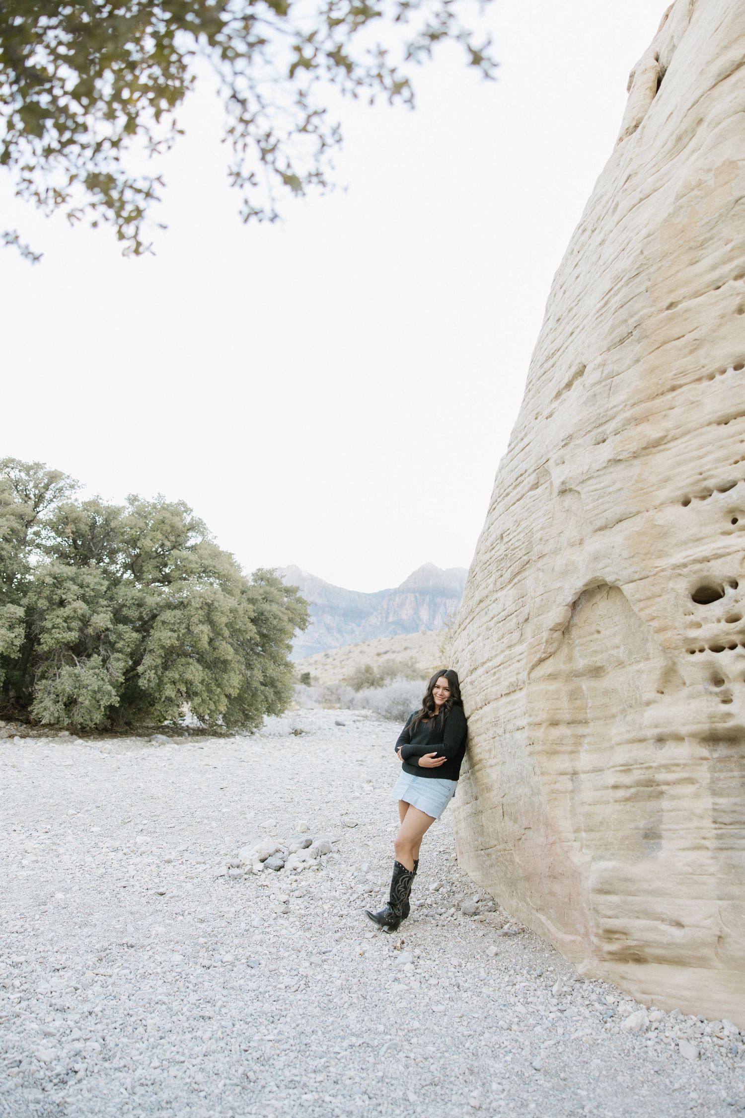 Someone poses against a white limestone rock wall in a desert landscape with mountains and trees in the background.