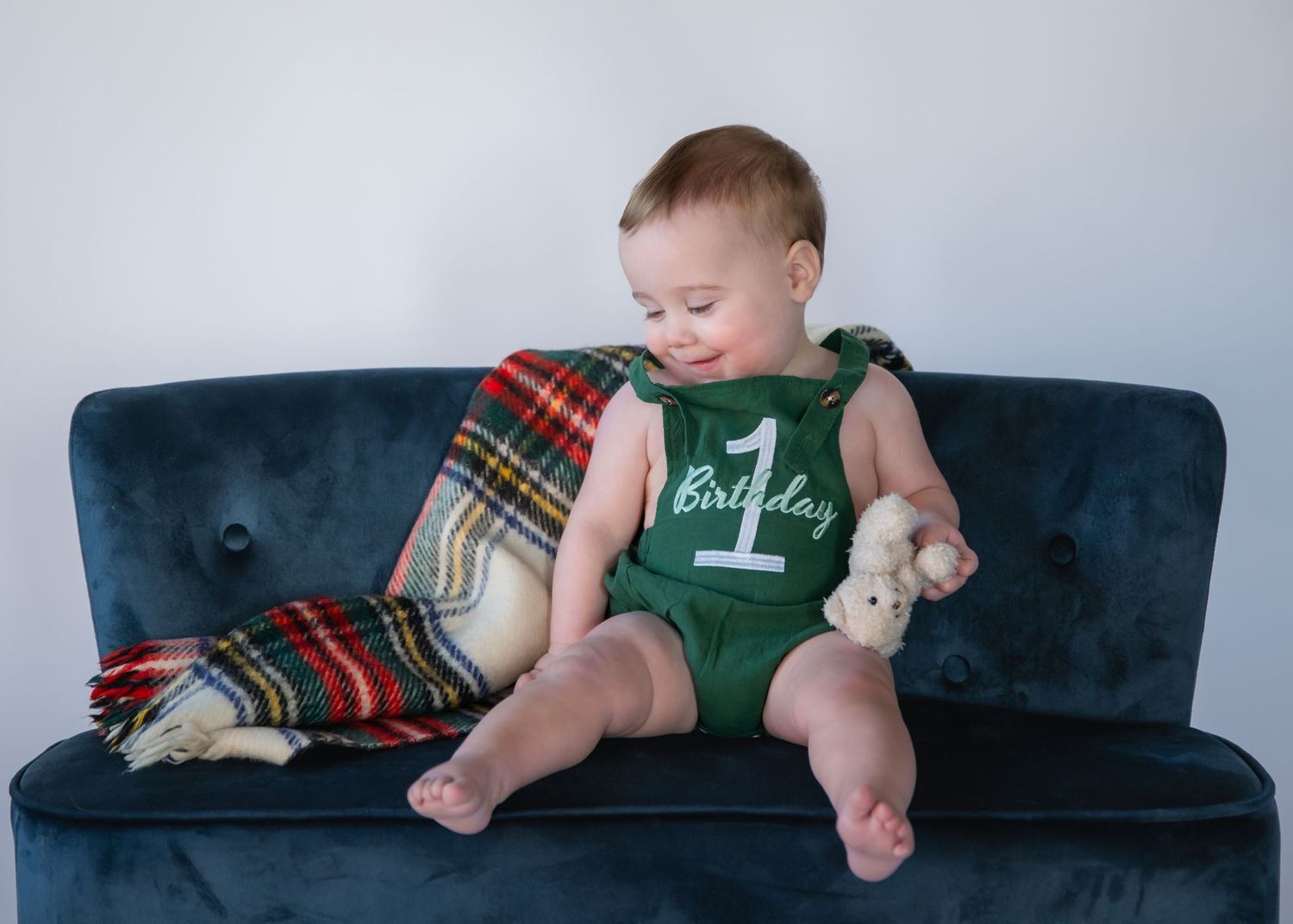 A baby in a green onesie sits on a blue velvet couch next to a plaid blanket and stuffed toy.