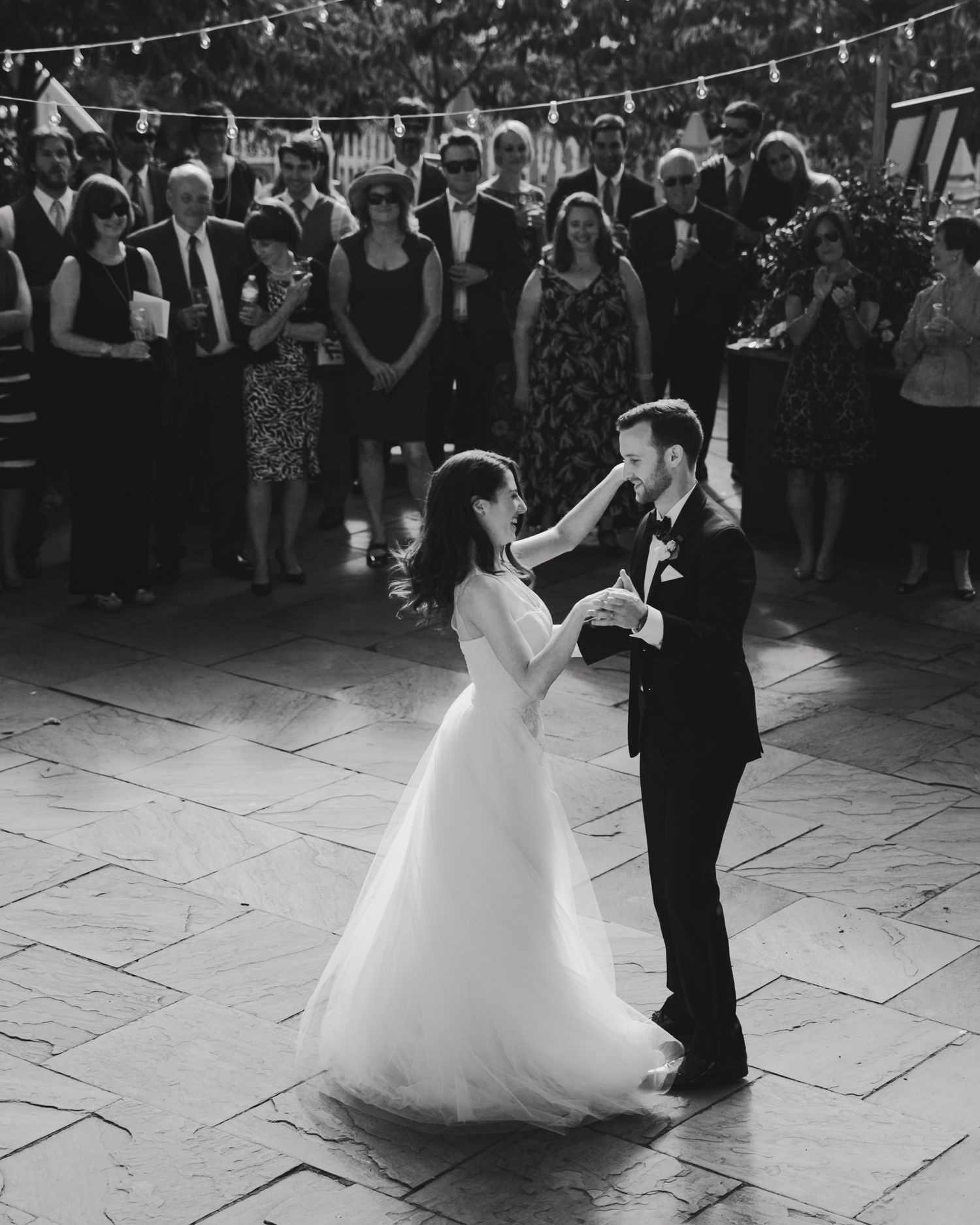 A couple shares their first dance at a wedding reception while guests watch from the background under string lights.