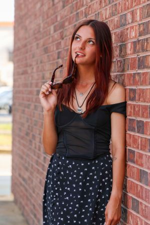 A young woman in a black top and floral skirt poses thoughtfully against a brick wall while holding sunglasses.