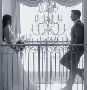 A romantic black and white photograph showing a couple on an elegant balcony with ornate chandeliers in the background.