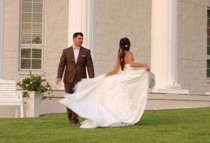 A couple in wedding attire walks on a manicured lawn in front of a white columned building at sunset.