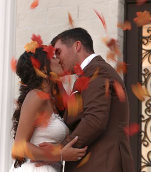 A couple embraces in a wedding kiss while orange autumn leaves fall around them.