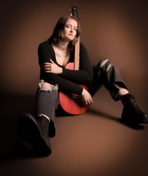 A moody portrait of a person in ripped jeans and boots posing with an acoustic guitar against a dark brown background.