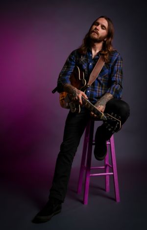 Musician in plaid shirt sits on purple stool playing acoustic guitar against dark pink studio backdrop.