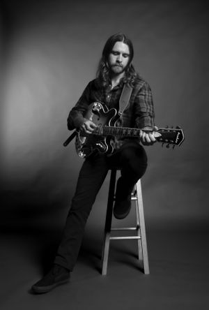 Black and white portrait of a musician seated on a stool playing an electric guitar, wearing plaid and denim.