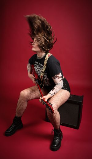 Rocker musician with flowing hair flips her head back while playing electric guitar on top of an amp on red backdrop.