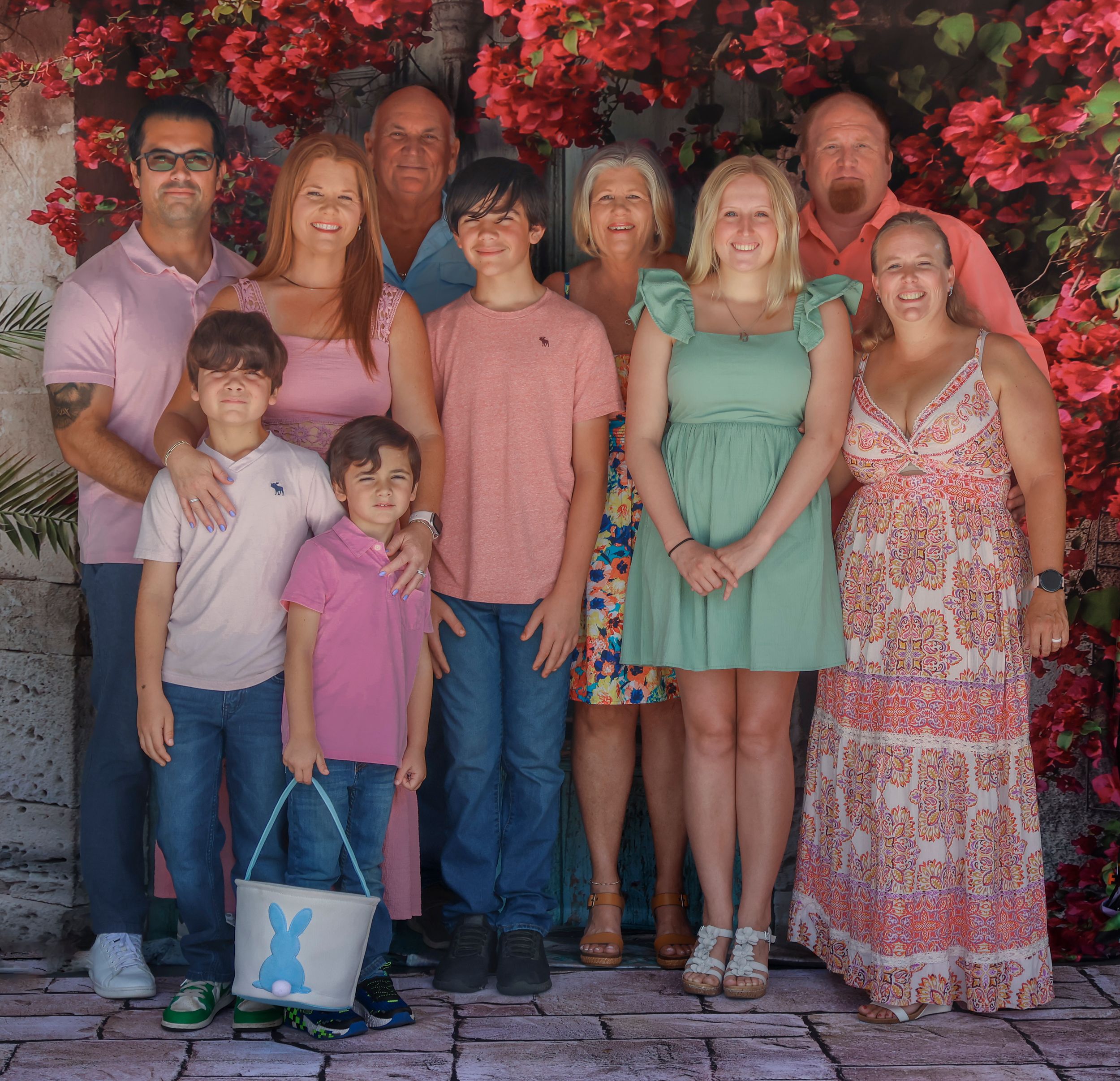 Family group, multigenerational portrait posing together outdoors against vibrant red bougainvillea flowers on stone wall background.