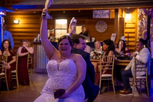A wedding couple enjoys a game of "who did first" in a rustic log cabin reception venue with warm lighting and guests seated around them.