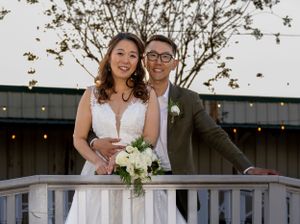 A couple poses together on a wooden deck railing under string lights during their wedding celebration.