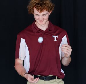 Student in maroon and white polo shirt juggles a golf ball against dark background.