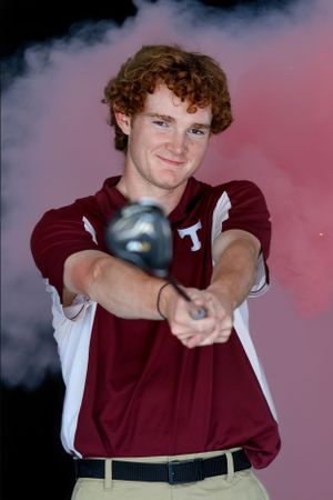 Student athlete in maroon and white uniform poses with a golf club in front of a misty pink background.