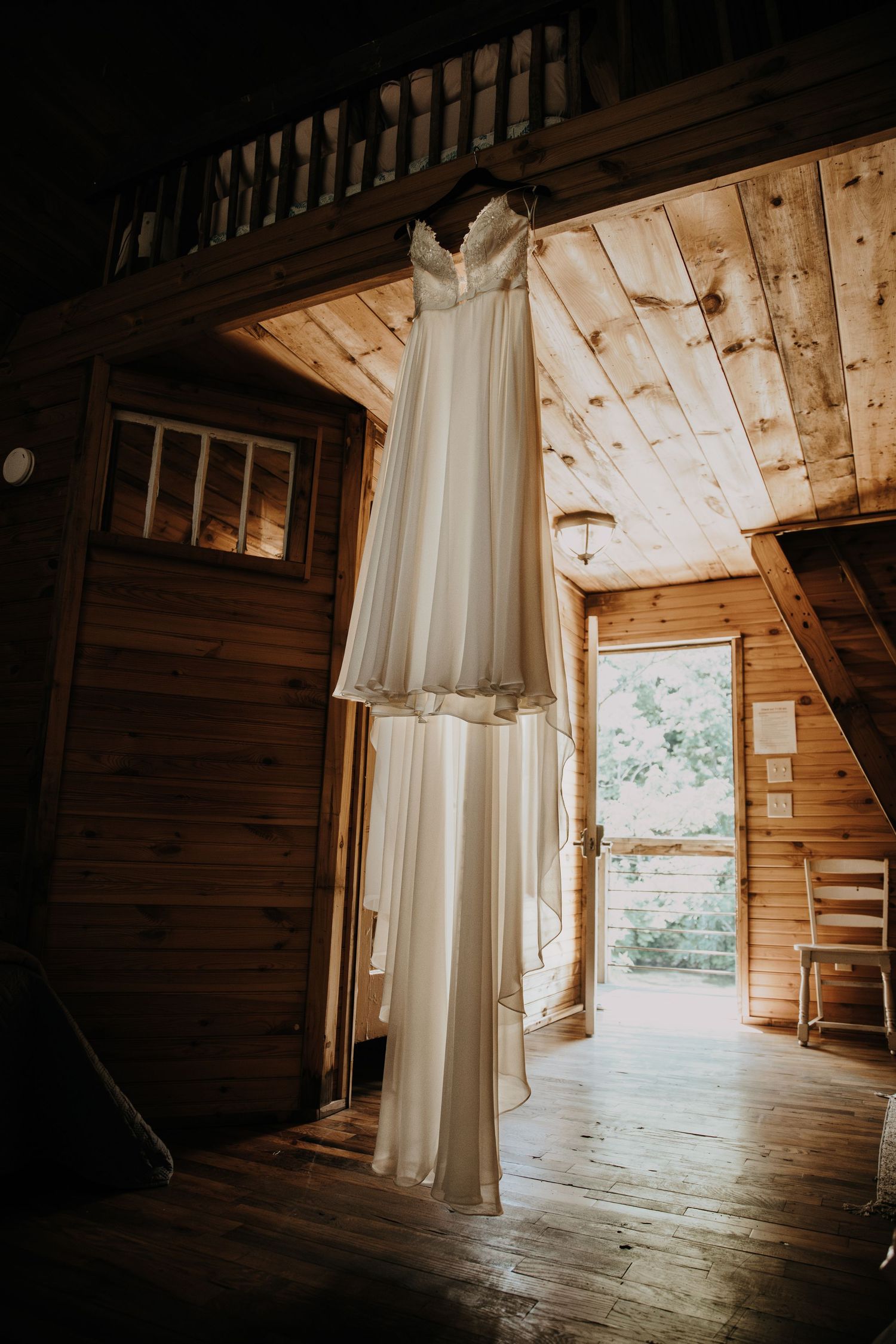 A white wedding dress hangs in rustic wooden cabin doorway with natural light streaming in.