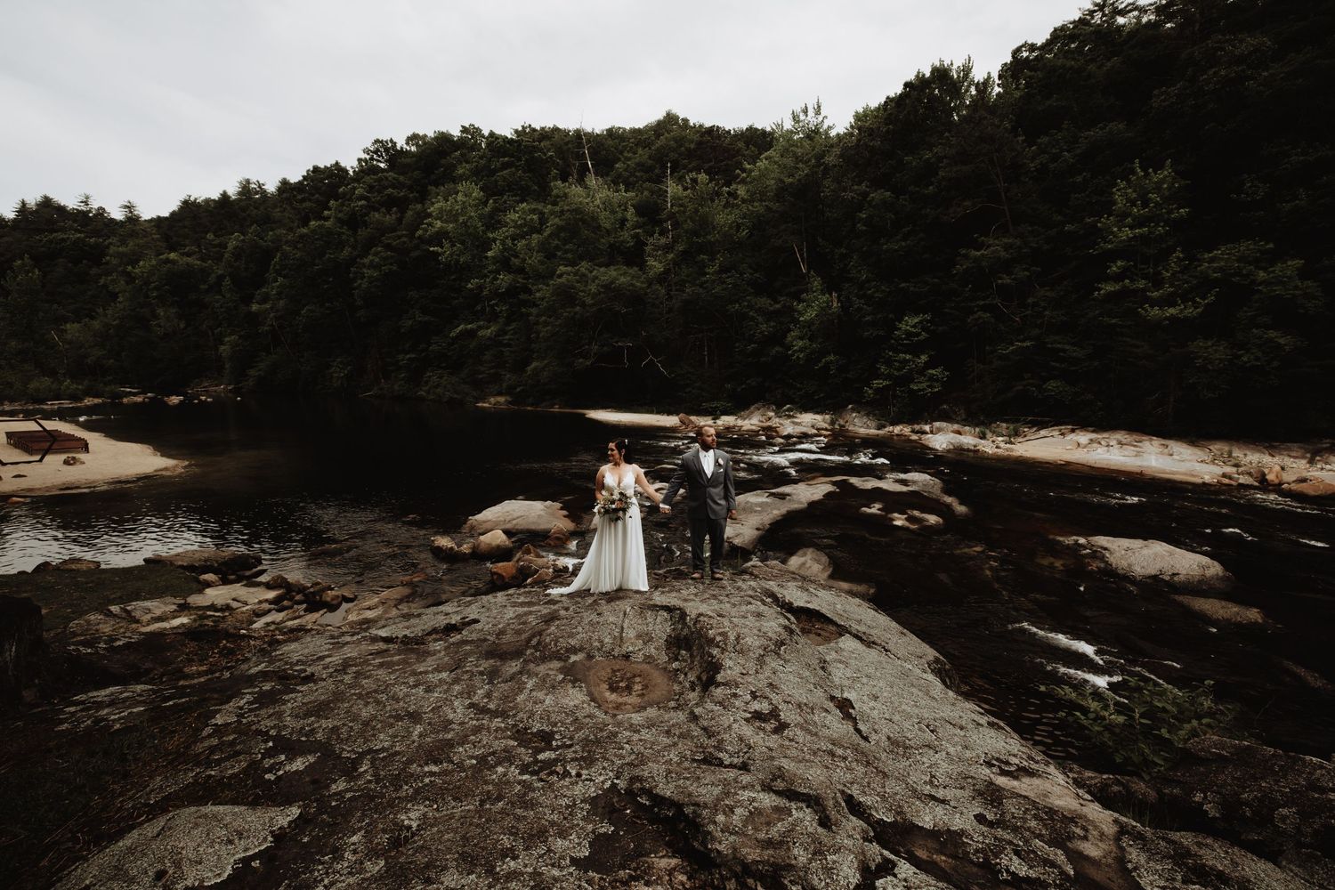 A dramatic wide-angle shot of newlyweds standing on rocky terrain surrounded by forest.