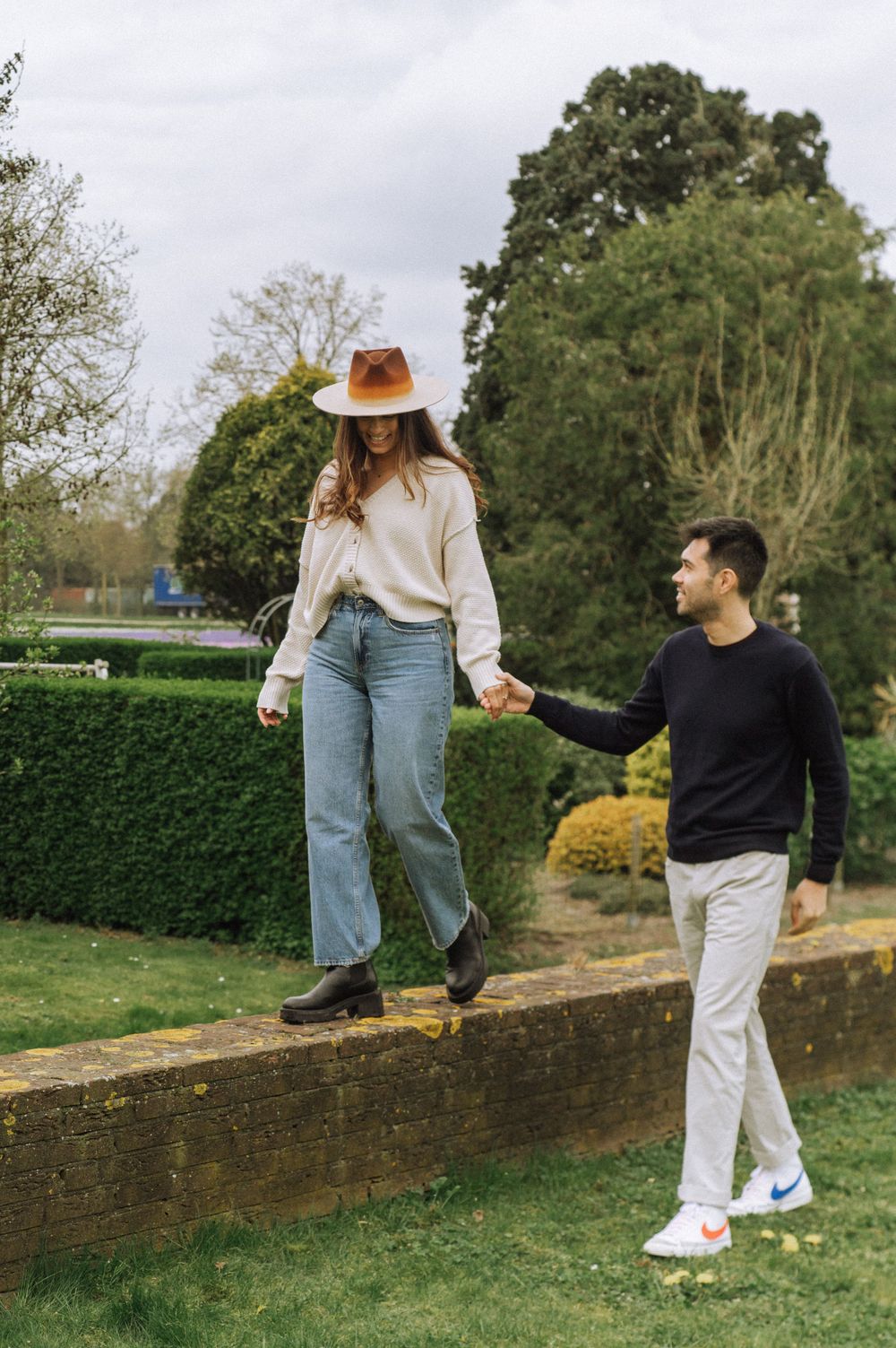 Couple playfully balancing on a stone wall in a garden setting.
