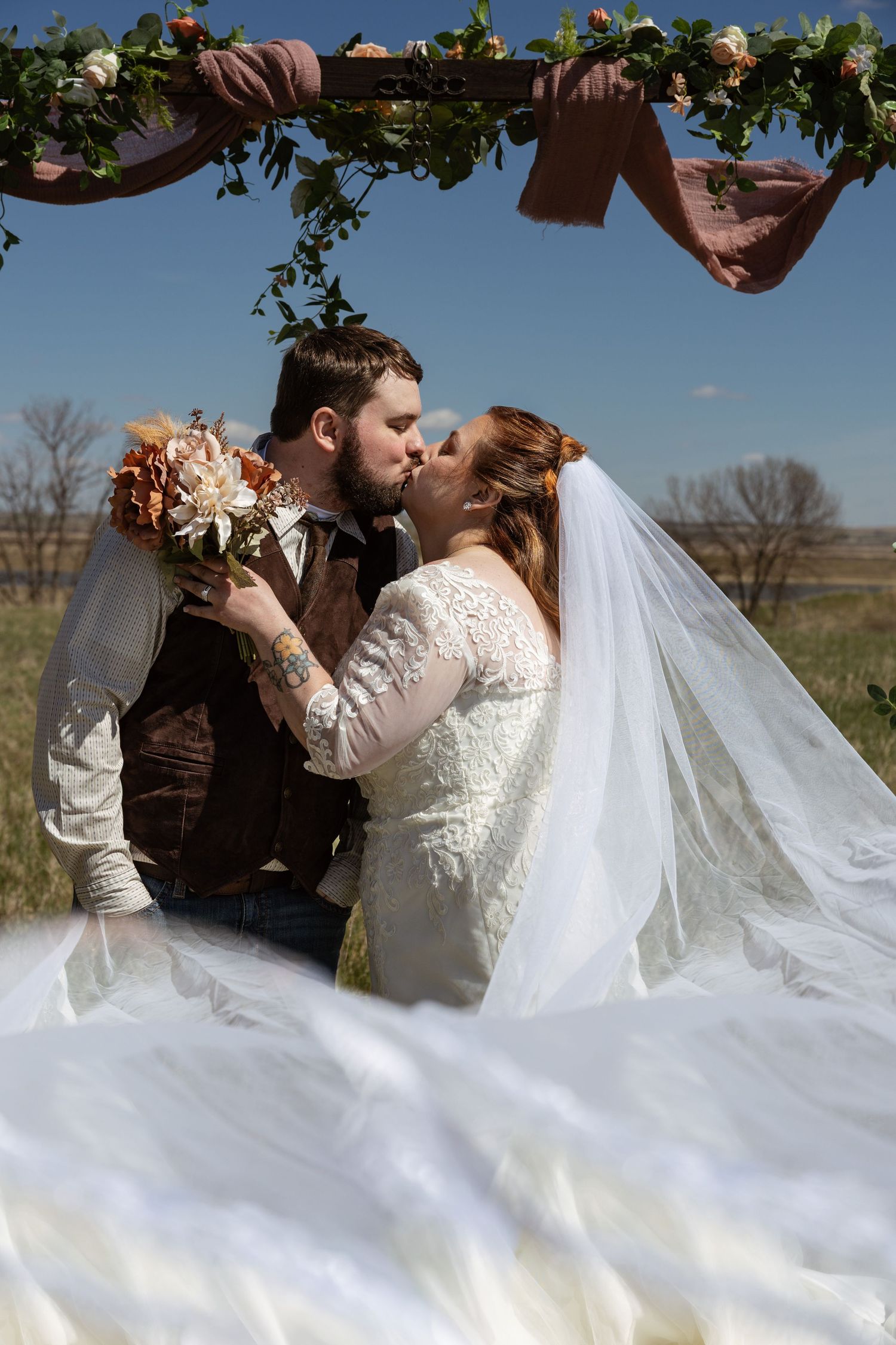 Bride and groom sharing a first kiss under a floral arch at an outdoor wedding.