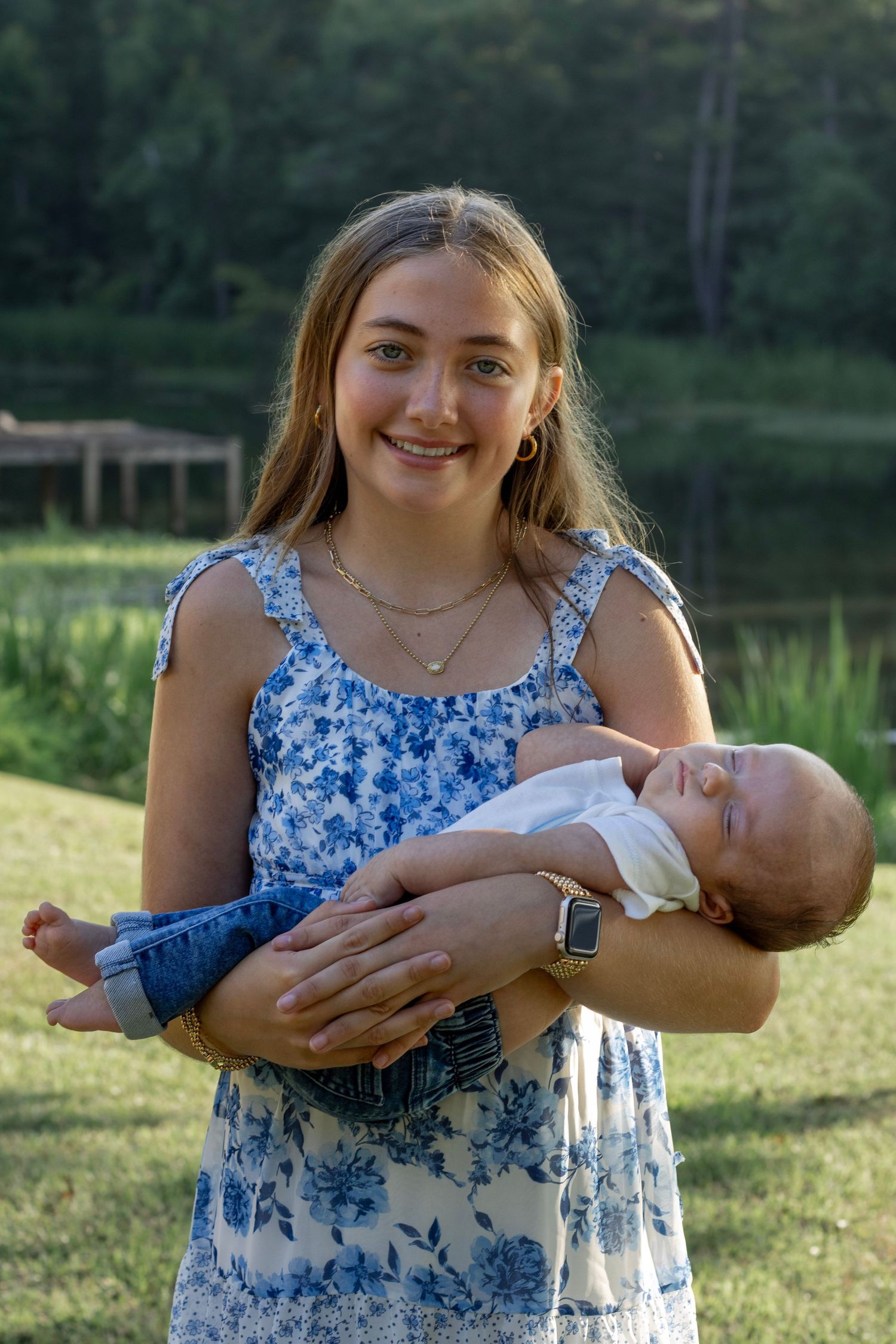 Big sister in a blue sundress holding her baby brother during outdoor photo shoot near a Lake Wateree, shoot by Raley and Roots Photography.