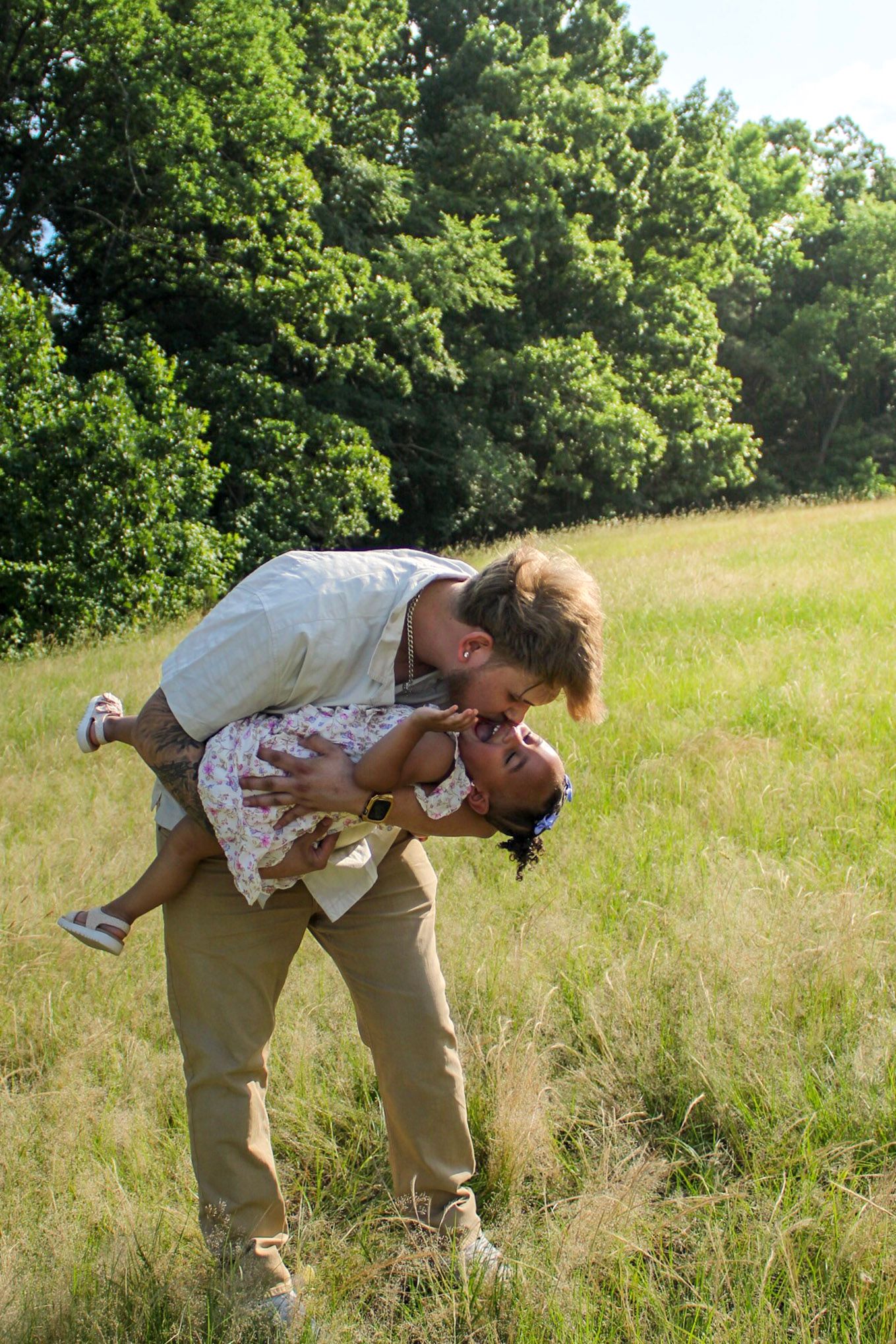 Raley and Roots Photography captures a tender moment between dad and daughter while embracing in a sunny field with trees in the background.