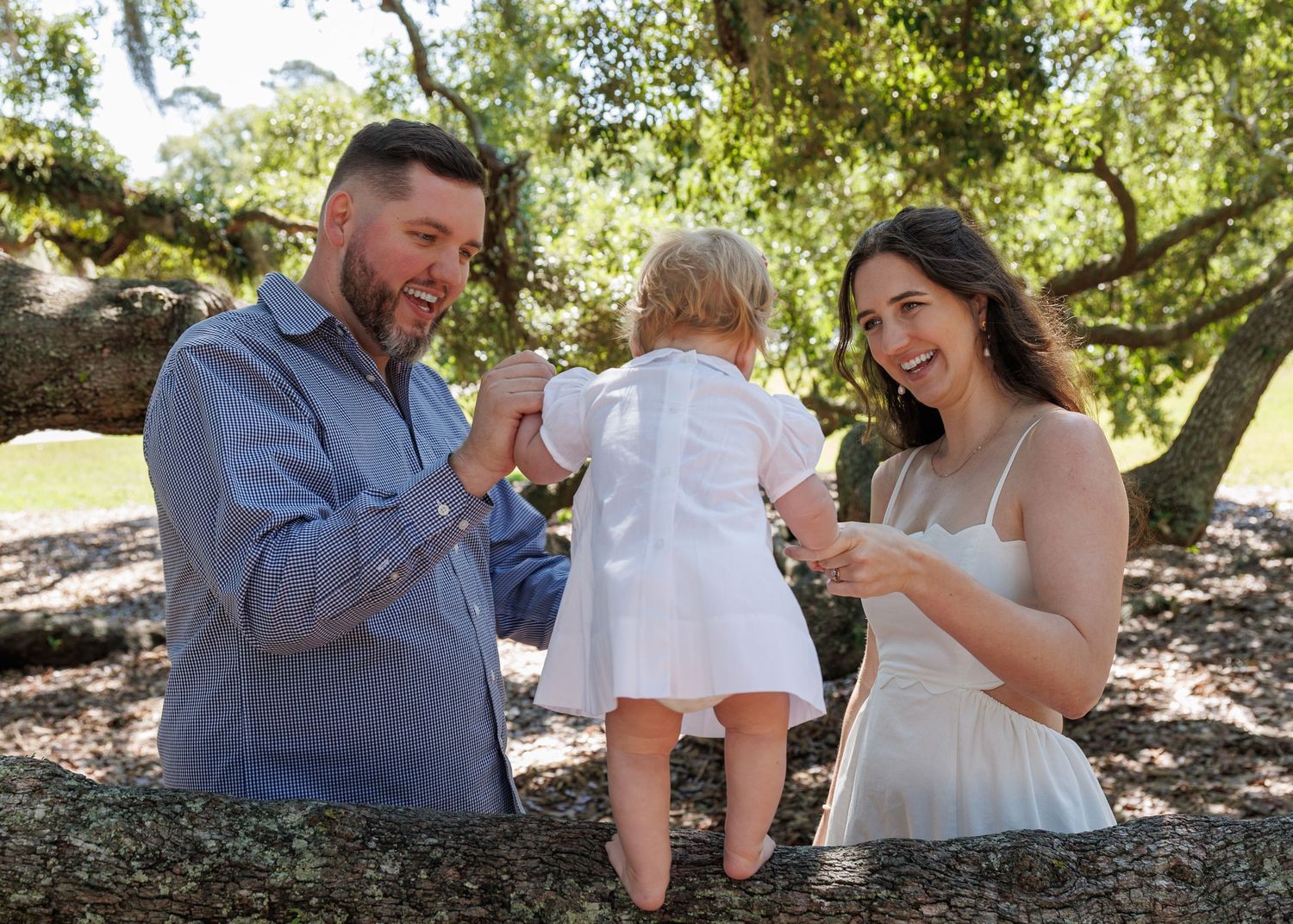 Happy family with parents helping toddler stand on a tree branch outdoors in a sunny park.