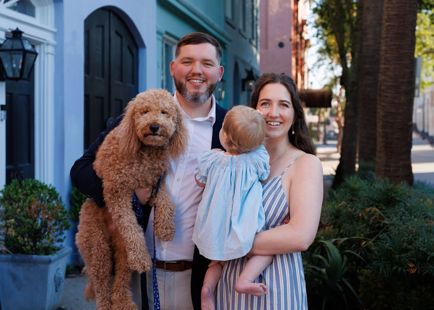 A smiling couple holds a curly golden doodle and a baby in a blue dress on a charming tree-lined street.