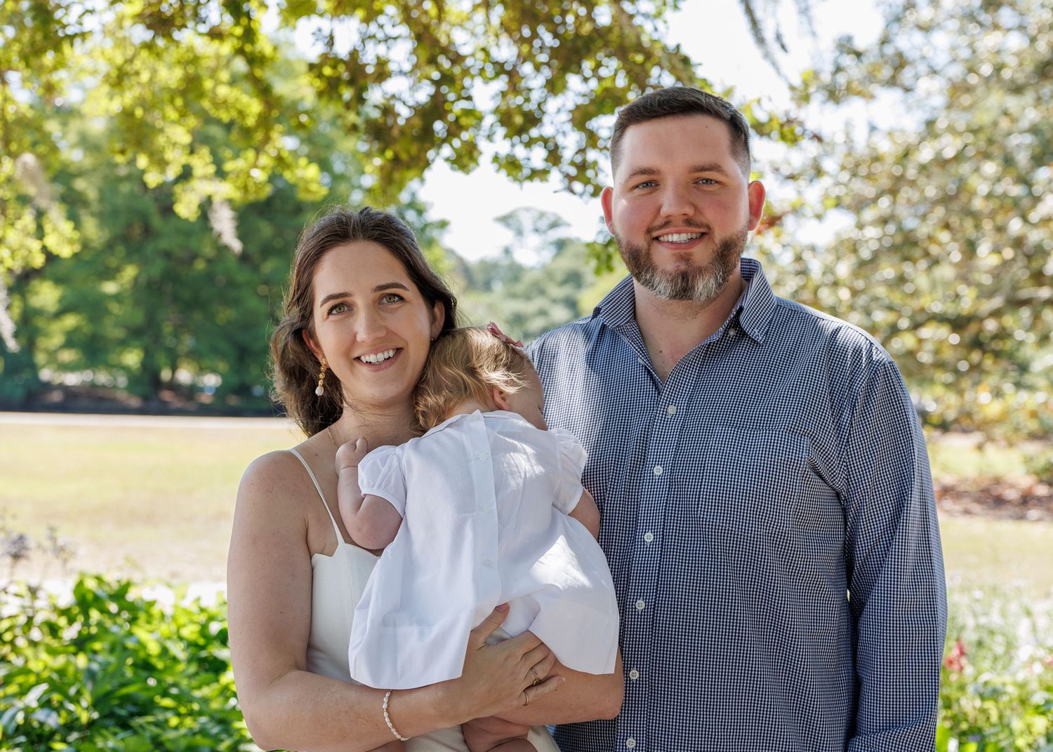 A smiling couple stands outdoors holding a newborn in a white gown, surrounded by lush green trees.