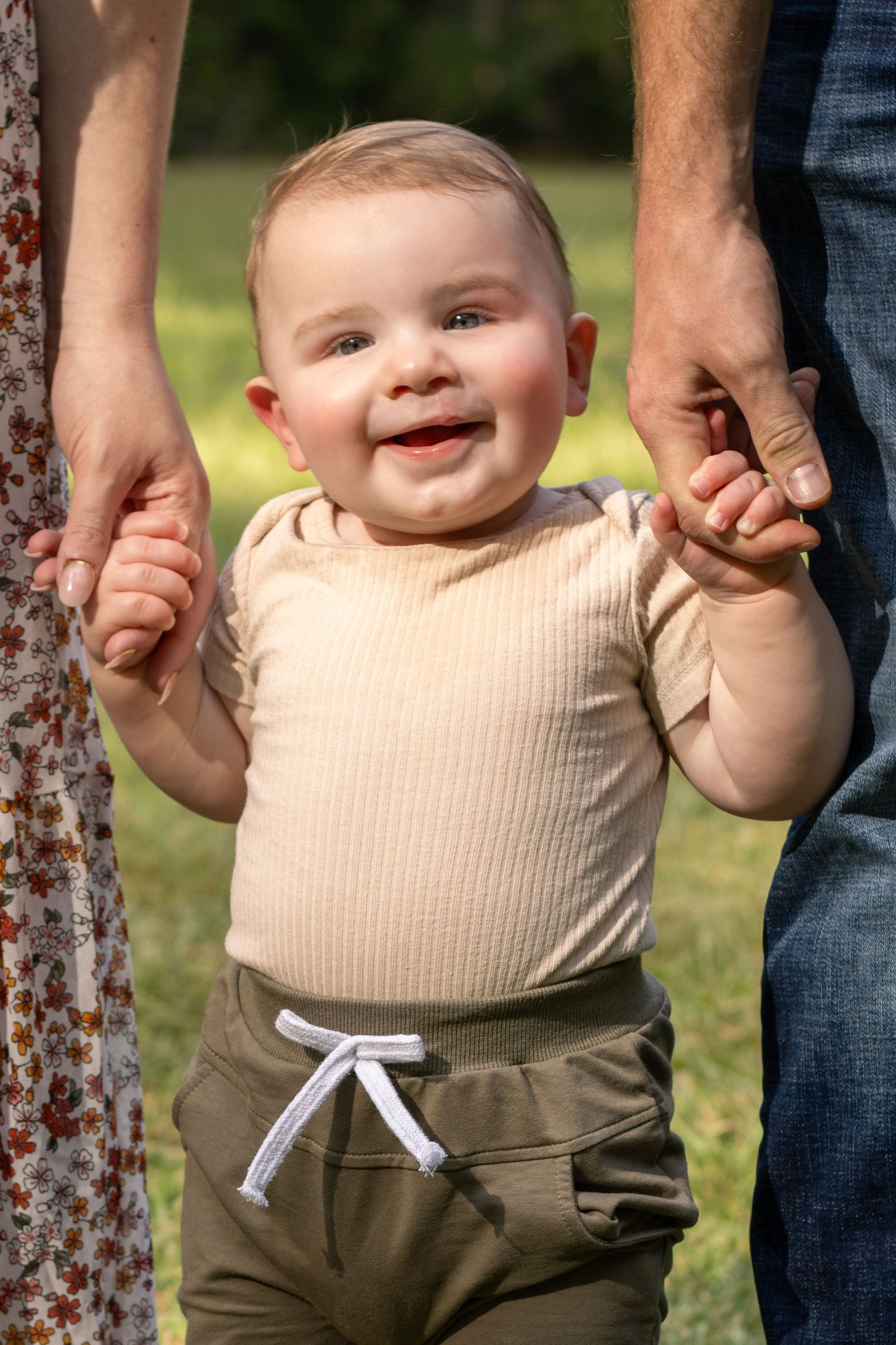 Close-up of a baby in beige outfit holding hands with parents outdoors.