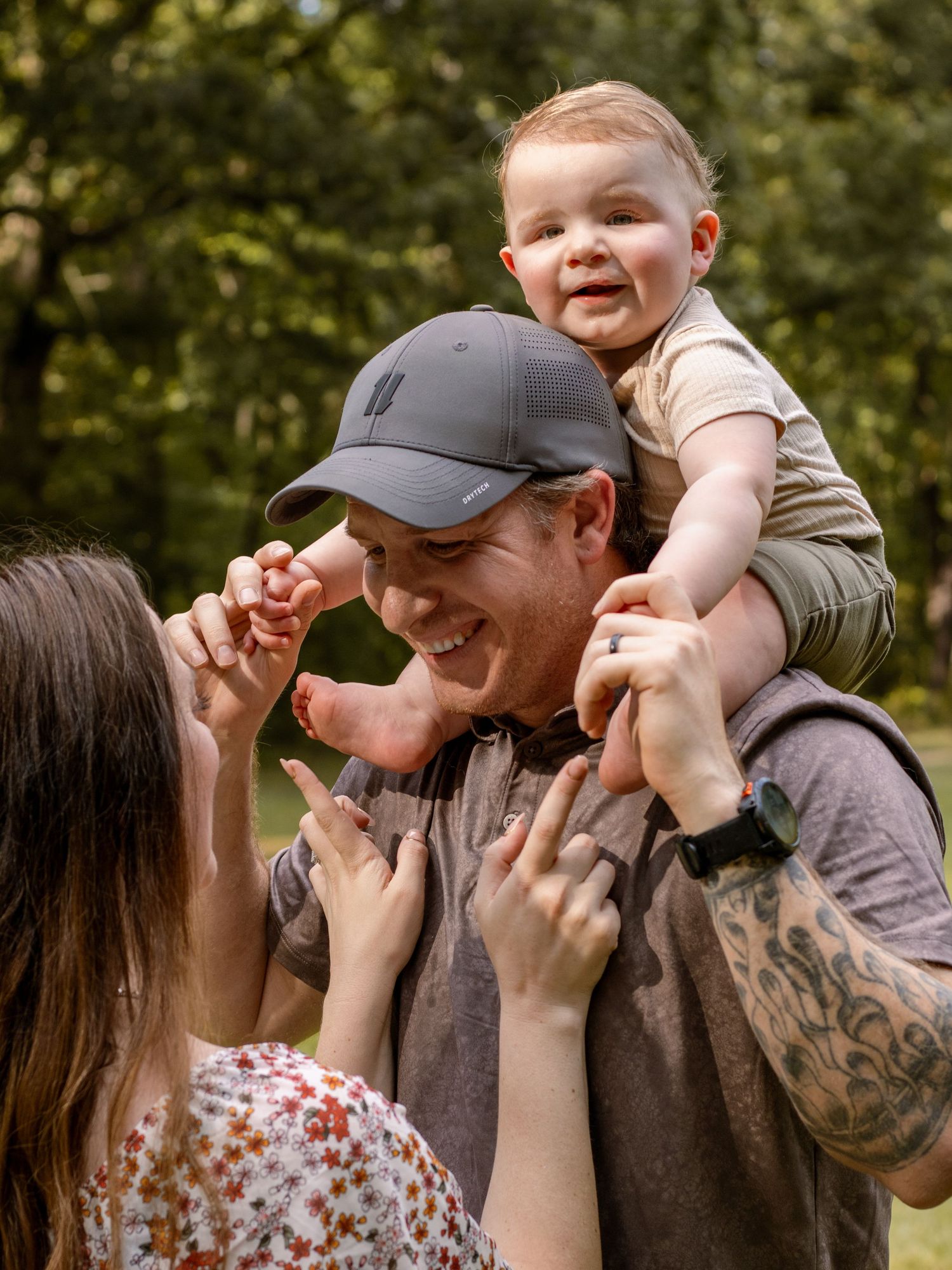 A happy father lifts a smiling baby on his shoulders while a woman reaches up joyfully outdoors.