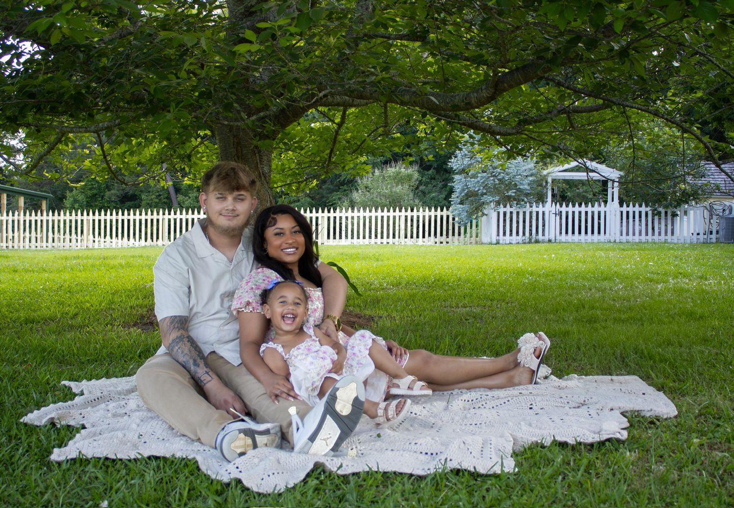 A family together on a white blanket in front of a white picket fence on a sunny summer day Ridgeway SC. Captured by Raley and Roots.