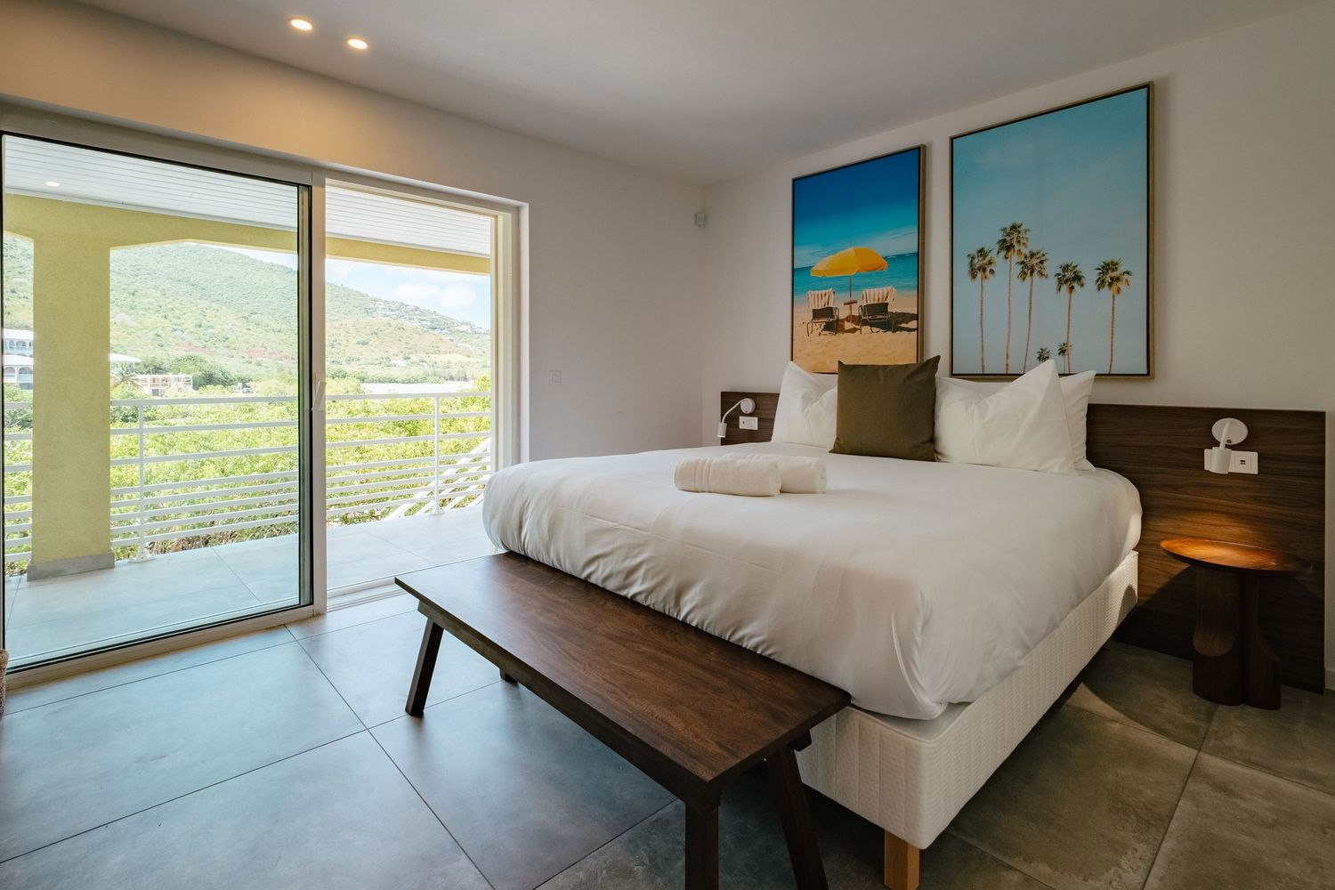 Bright hotel room with white bedding, palm tree artwork, and large sliding glass doors overlooking mountain views.
