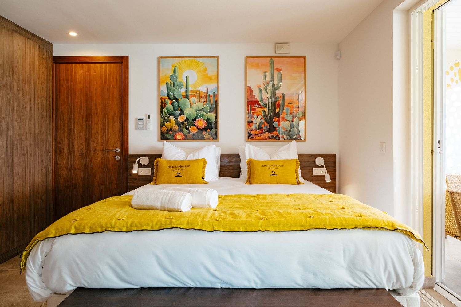 Modern bedroom with white bed featuring yellow bedding and two colorful cactus artwork prints on the wall.